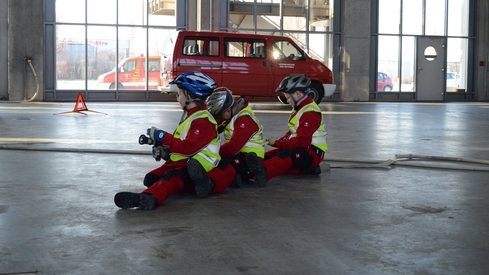 drei hintereinander auf dem Boden sitzende Kinder die einen Löschschlauch halten