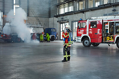 ABC-Übung in der Feuerwehrübungshalle. Im Vordegrund der Einsatzleiter am Handsprechfunkgerät, daneben ein Löschfahrzeug. Im Hintergrund Einsatzkräfte in Schutzanzügen bei 