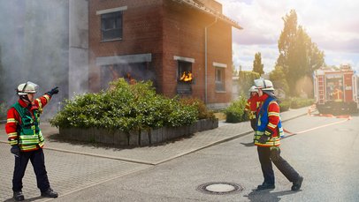 Drei Einsatzkräfte bei einer Übung vor dem Feuerwehr-Übungshaus. Im Hintergrund ein Löschfahrzeug mit einer weiteren Einsatzkraft