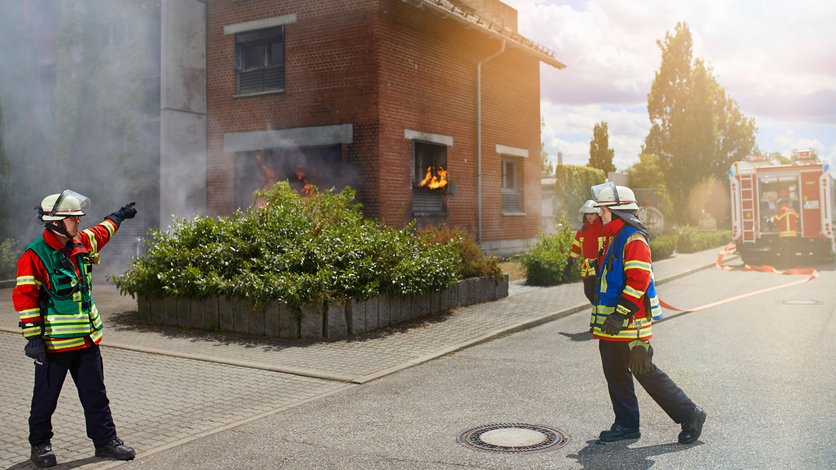 Drei Einsatzkräfte bei einer Übung vor dem Feuerwehr-Übungshaus. Im Hintergrund ein Löschfahrzeug mit einer weiteren Einsatzkraft
