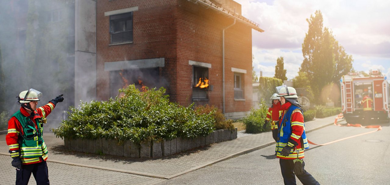 Drei Einsatzkräfte bei einer Übung vor dem Feuerwehr-Übungshaus. Im Hintergrund ein Löschfahrzeug mit einer weiteren Einsatzkraft