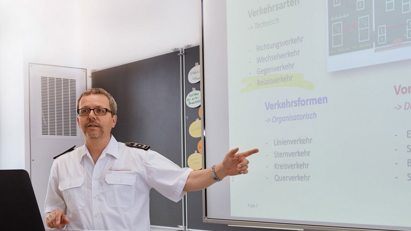 Ein Ausbilder steht vor einem Multifunktionsboard auf dem eine Bild einer Powerpointfolie eingeblendet ist