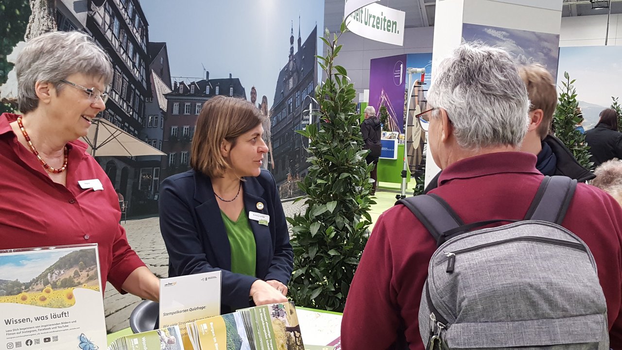 Zwei Frauen stehen hinter einem Messestand des Biosphärengebiets auf der CMT in Stuttgart. Zwei interessierte Personen informieren sich.