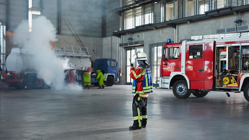 ABC-Übung in der Feuerwehrübungshalle. Im Vordegrund der Einsatzleiter am Handsprechfunkgerät, daneben ein Löschfahrzeug. Im Hintergrund Einsatzkräfte in Schutzanzügen bei 