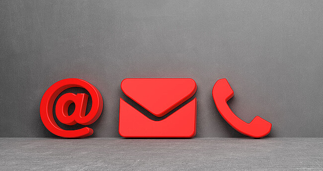 Ein rotes @-Zeichen, ein Briefumschlag und ein Telefonhörer als Symbol vor einer grauen Wand