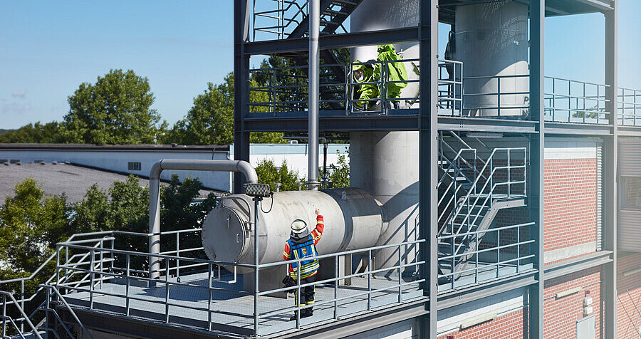 ABC-Übung auf der Chemie-Übungsanlage mit zwei Einsatzkräften in Schutzausrüstung auf einem Tankturm und dem Einsatzleiter auf der Ebene darunter.